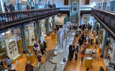View of a museum hall from an upstairs gallery, showing groups of people looking at exhibits.